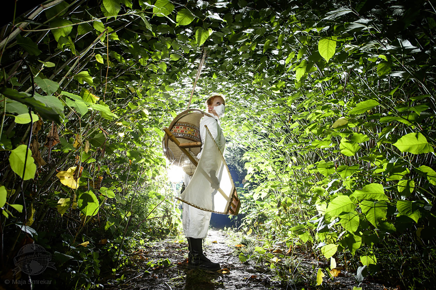 Femme en blanc portant un kit de survie à l'anthropocène dans un tunnel végétal.